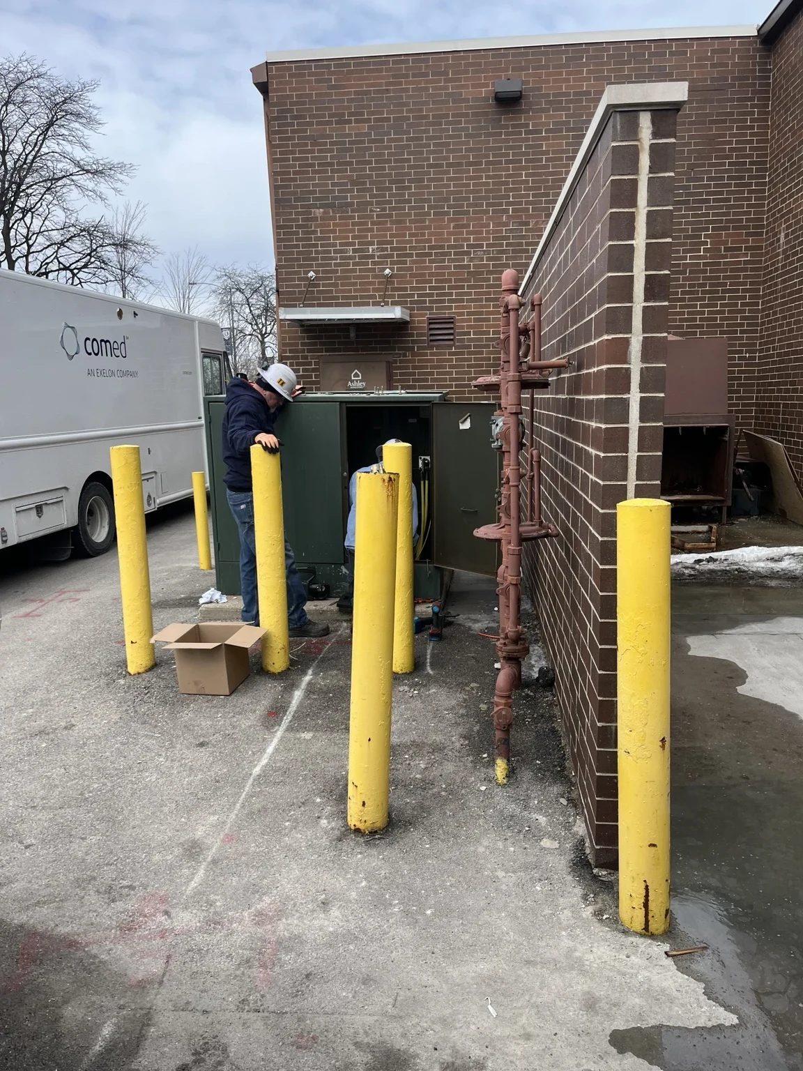 Exterior electrical equipment beside a brick wall and protective yellow bollards.