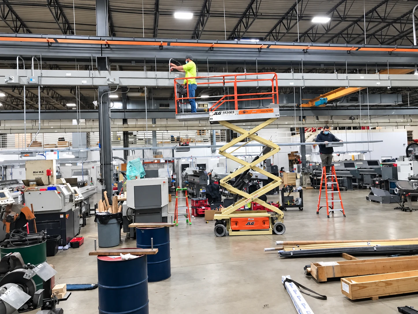 Electricians working from a scissor lift and ladder on overhead electrical installation in the machine shop.
