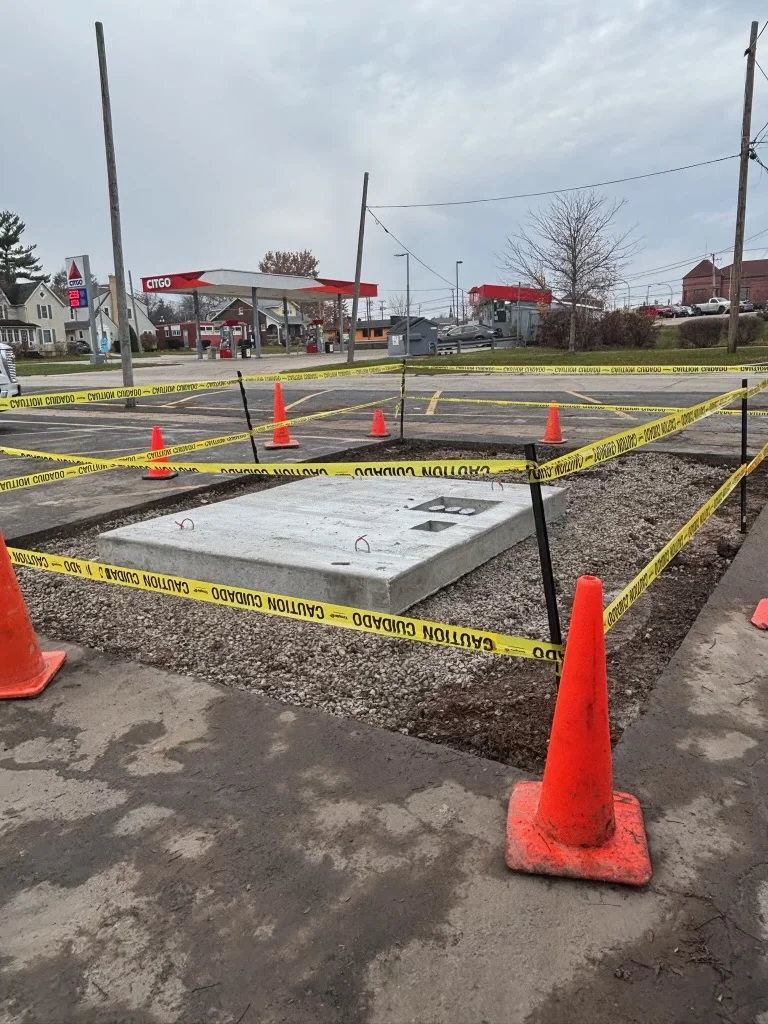 Precast concrete transformer pad surrounded by gravel, caution tape, and traffic cones.