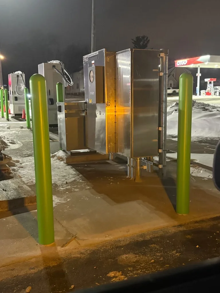 Electrical metering enclosure and green bollards at the charging site with snow on the ground.