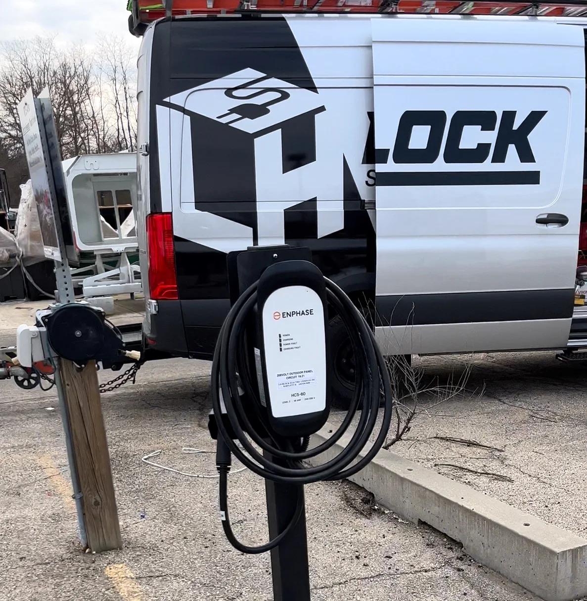 Outdoor pedestal-mounted Enphase charger on a concrete base with an LH Block service van behind it.
