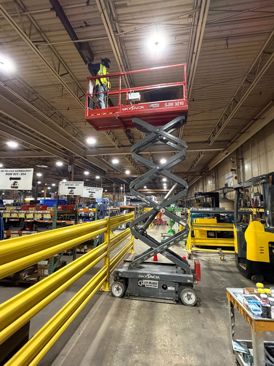 LH Block electrician working from a scissor lift above a manufacturing aisle beside yellow guardrails.